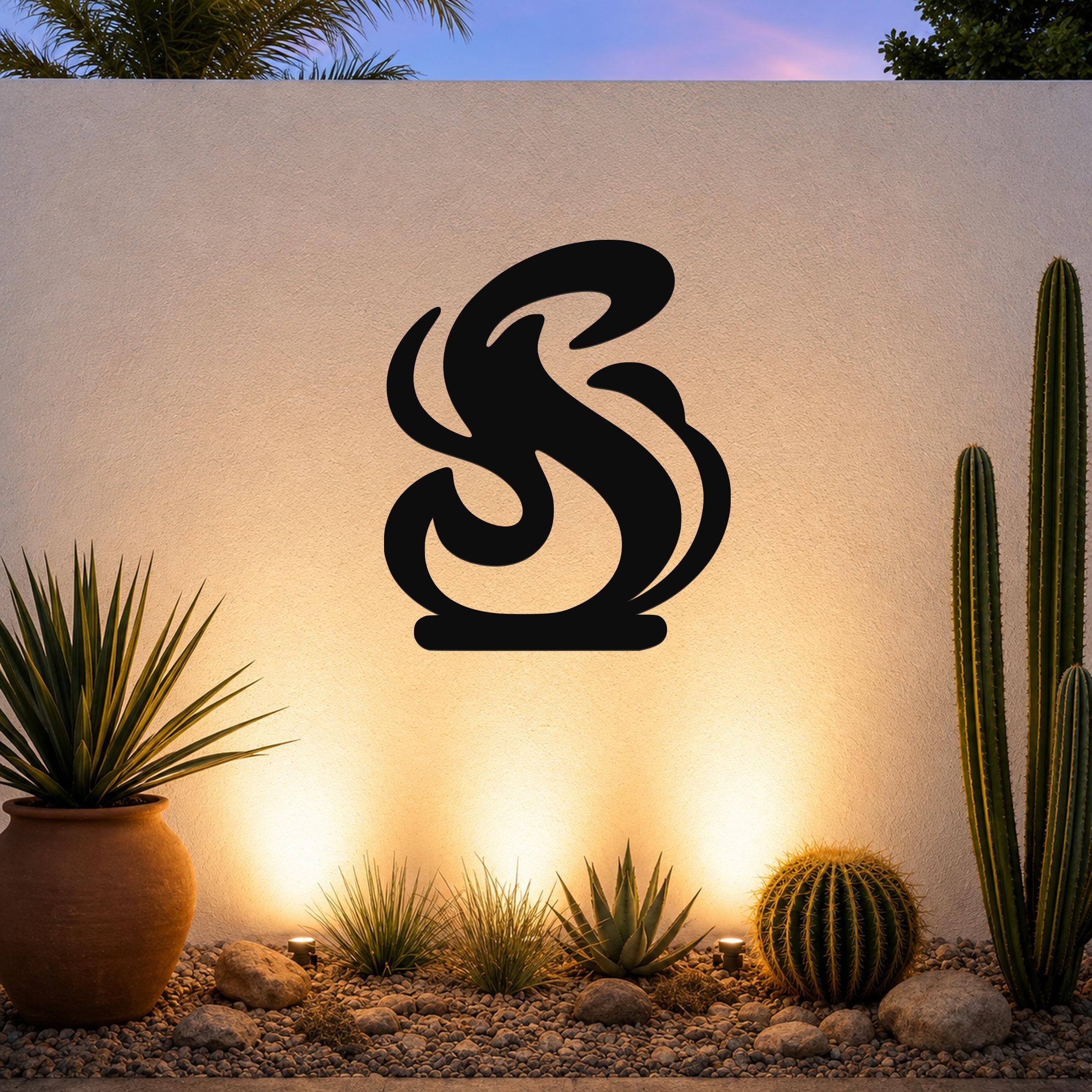 Black metal flame silhouette wall art with flowing curves, mounted on an outdoor wall above a desert garden with cacti and warm uplighting.