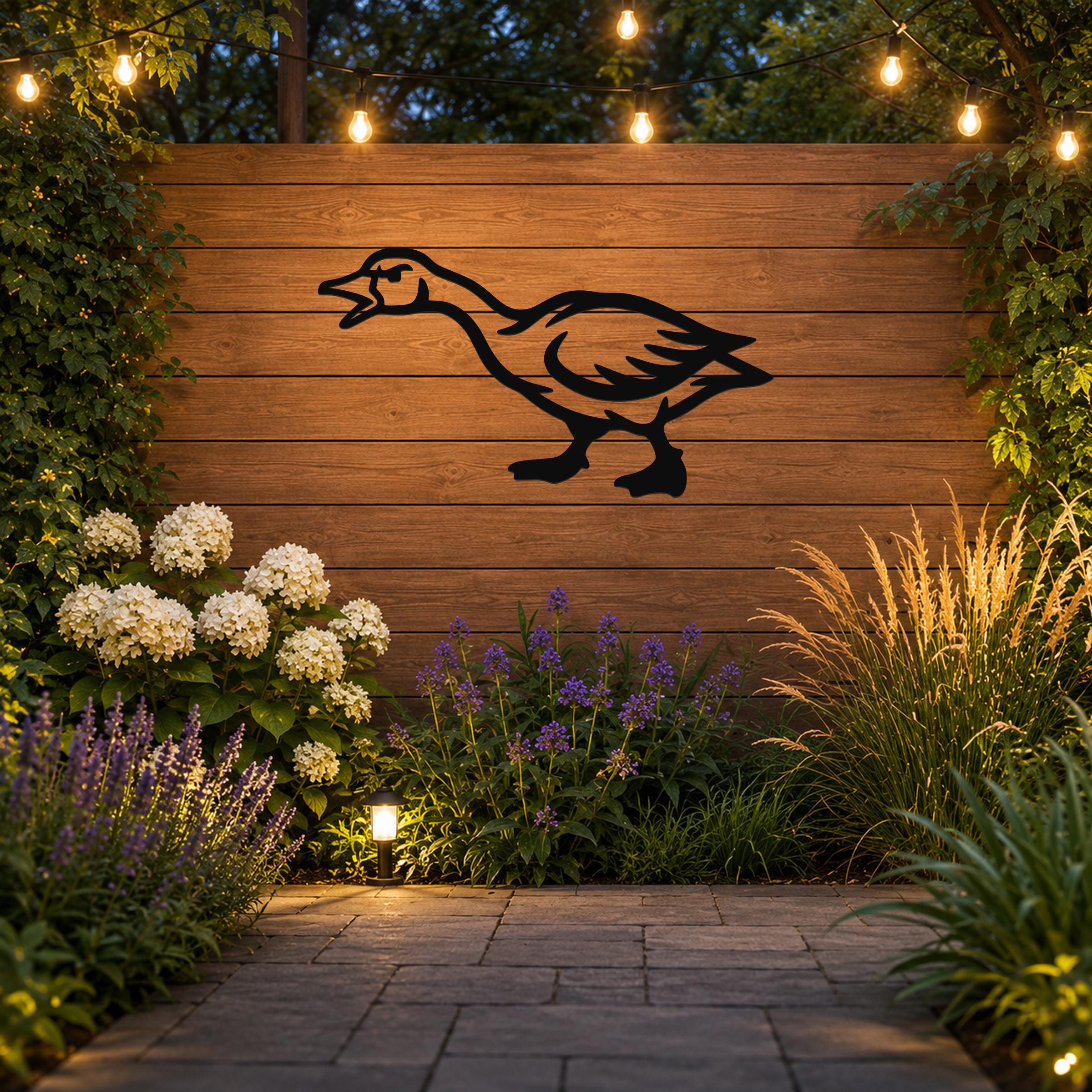 Black metal goose wall art silhouette mounted on a wooden fence in a garden, illuminated by string lights and surrounded by flowers and plants.