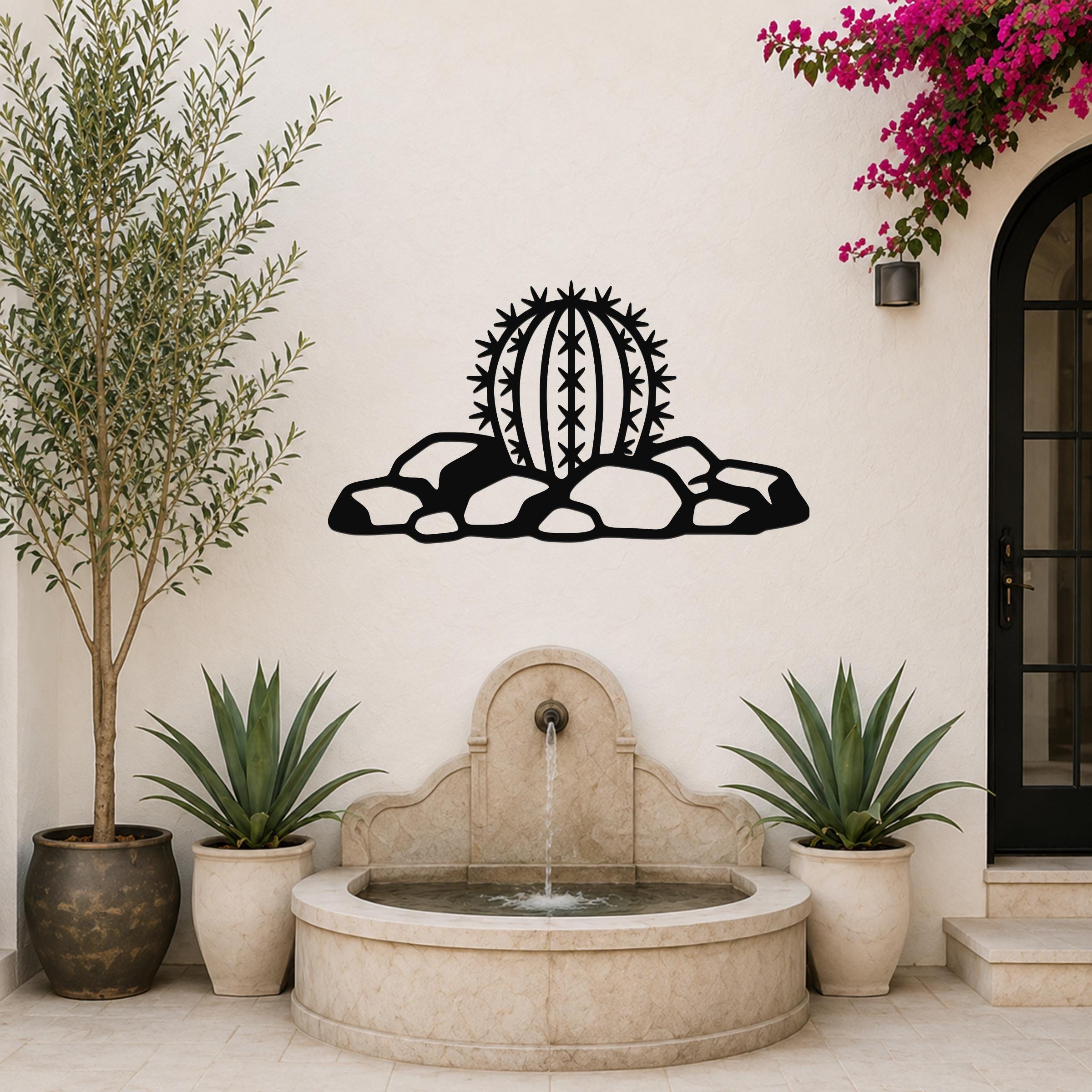 Black metal barrel cactus wall art with spines and rocks cutout, mounted on a white outdoor wall above a stone fountain.