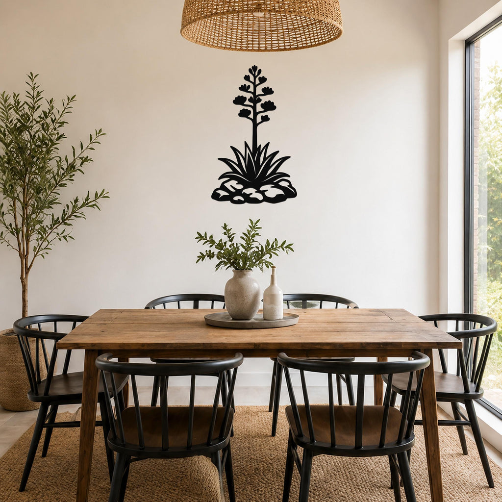Black metal agave wall sculpture with blooming flower and leaf cutouts, mounted on a white wall above a wooden dining table in a bright dining room.