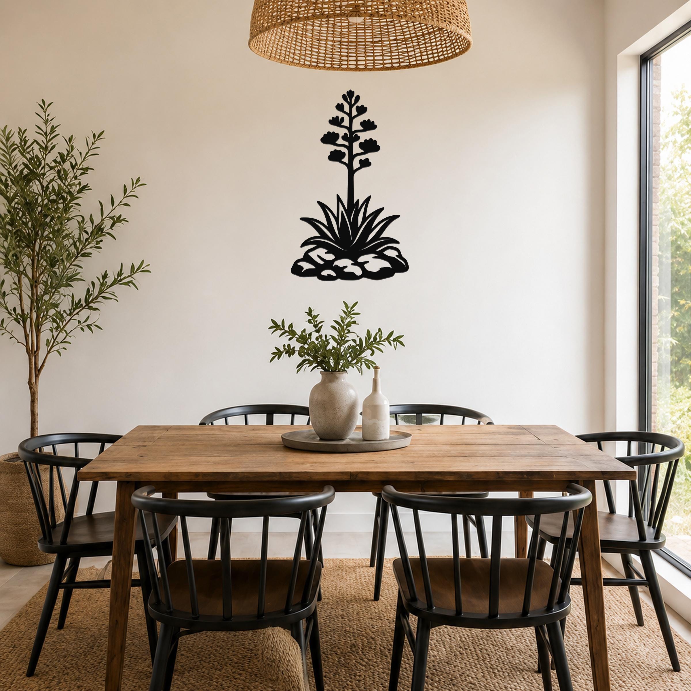 Black metal agave wall sculpture with blooming flower and leaf cutouts, mounted on a white wall above a wooden dining table in a bright dining room.
