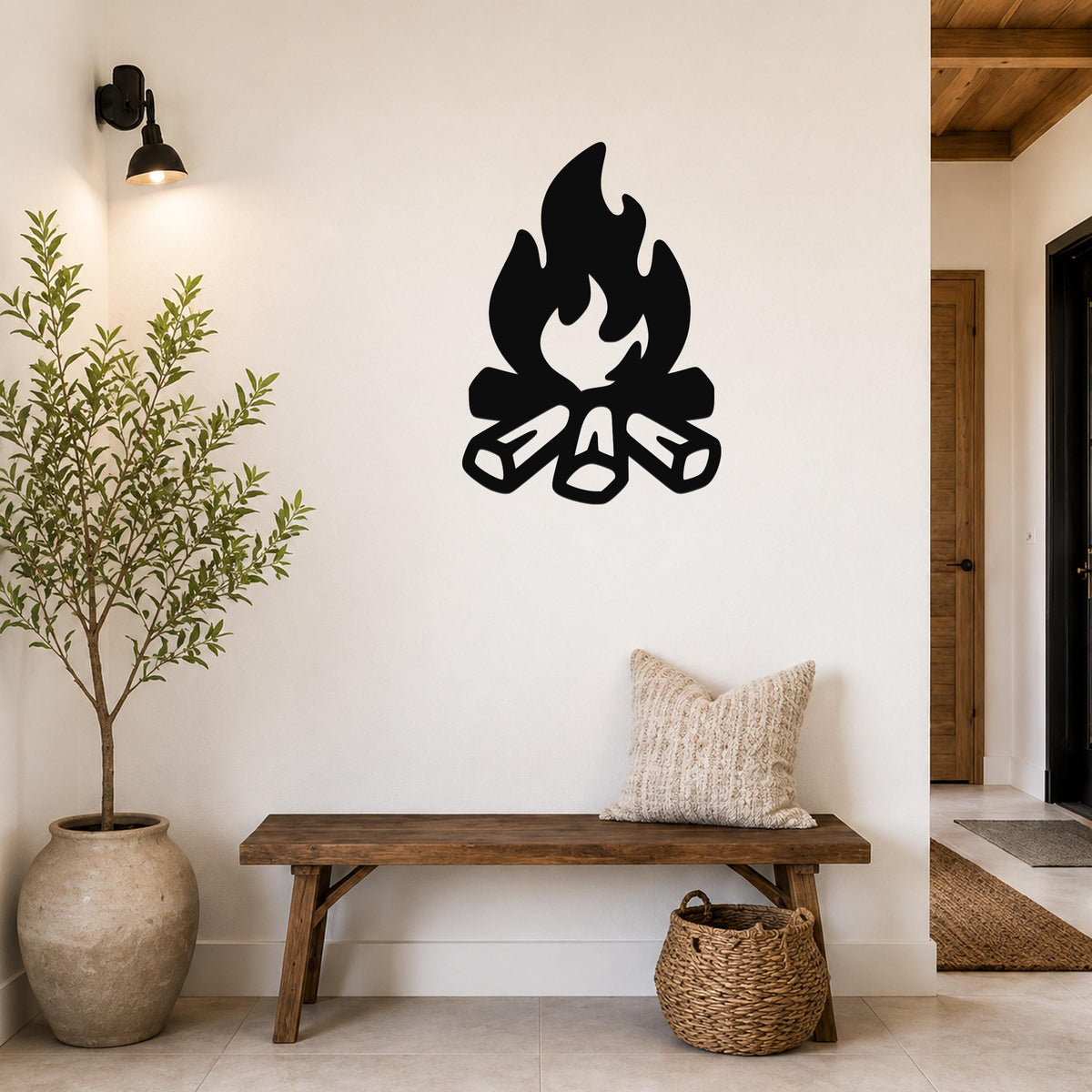 Black metal campfire wall art with flame and log silhouette, mounted on a white wall above a wooden bench in a rustic entryway.