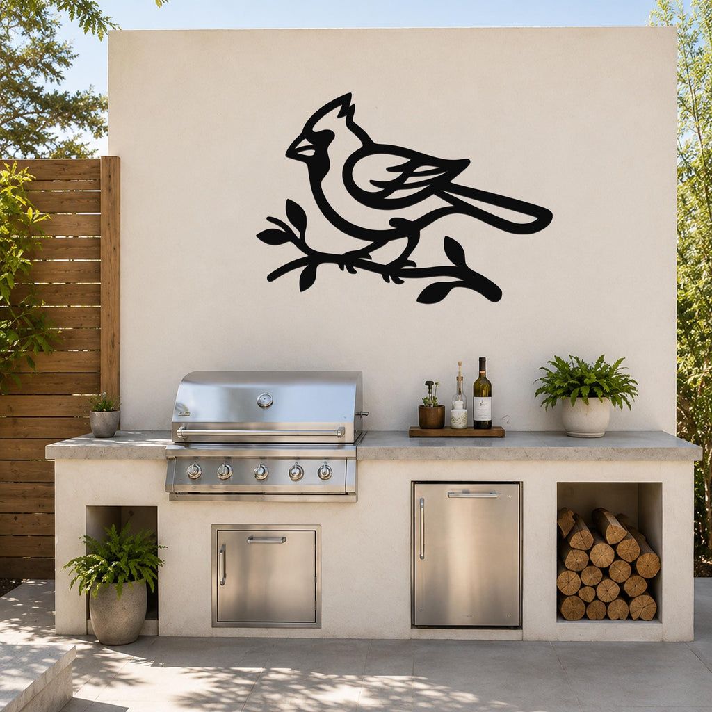 Black metal cardinal wall art in silhouette perched on a branch, mounted on an outdoor kitchen wall above a stainless steel grill.
