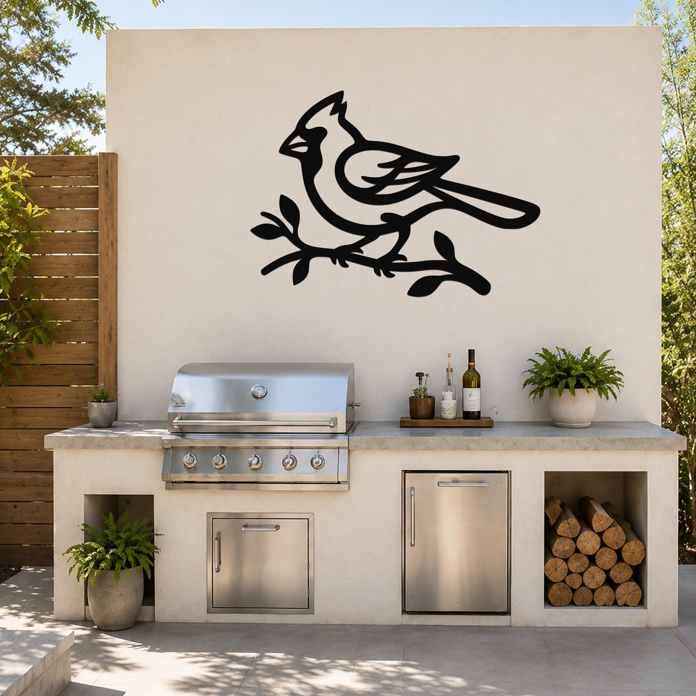 Black metal cardinal wall art in silhouette perched on a branch, mounted on an outdoor kitchen wall above a stainless steel grill.