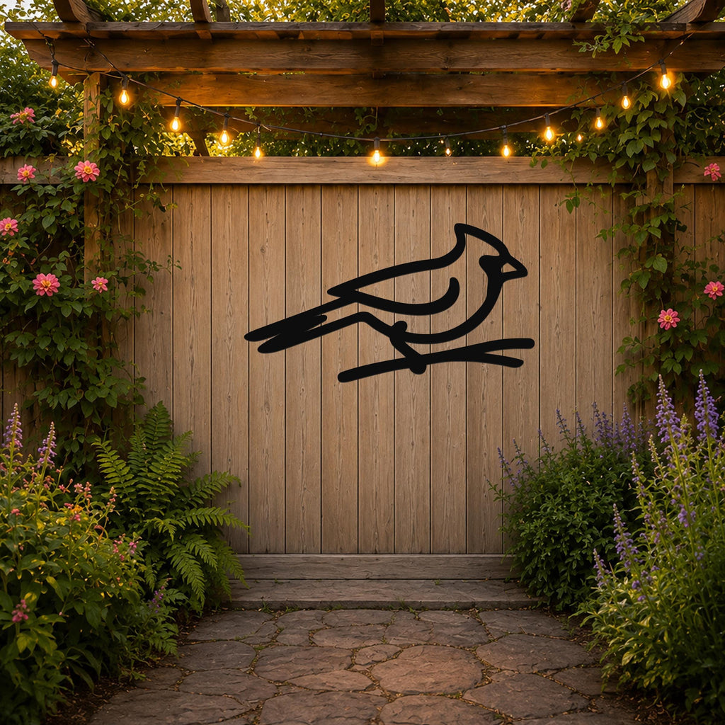 Black metal cardinal wall art silhouette perched on a branch, mounted on a wooden fence in a garden with string lights and greenery.