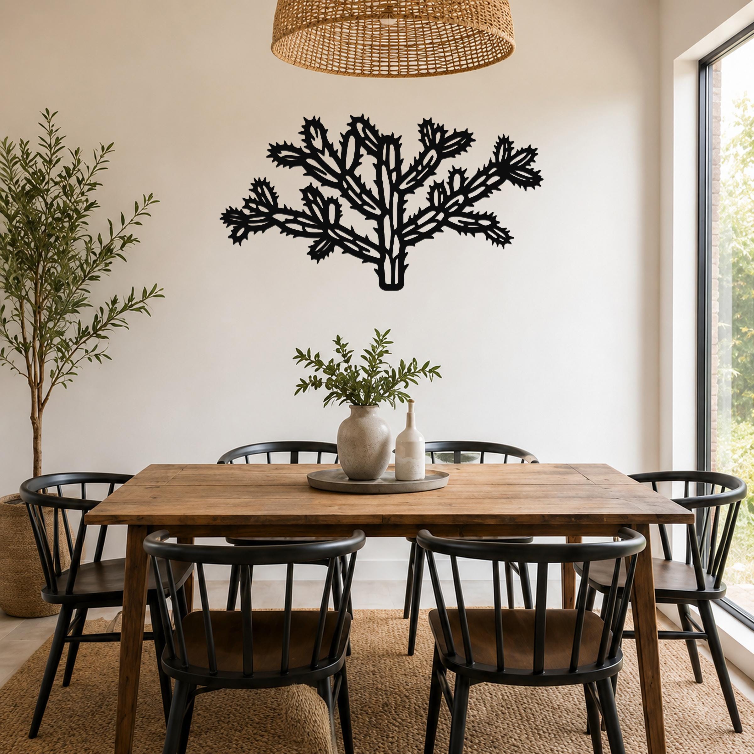 Black metal cholla cactus wall art with intricate branch cutouts, mounted on a white wall above a wooden dining table in a bright room.