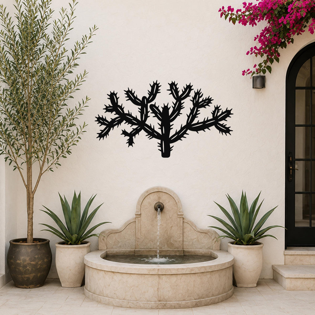 Black metal Cholla cactus wall art with textured spine cutouts, mounted on a white outdoor wall above a stone fountain.