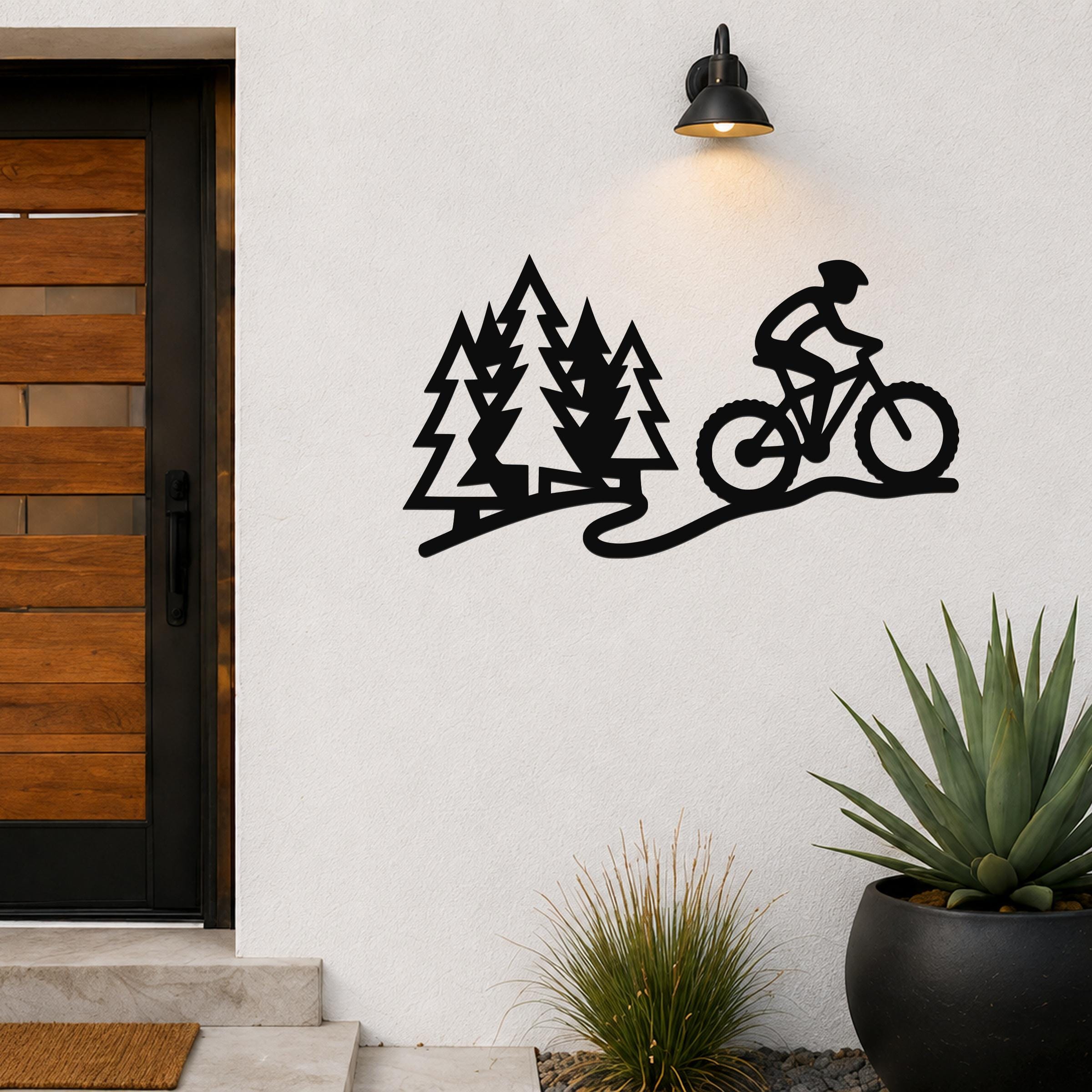 Black metal wall art cutout of a cyclist on a trail with pine trees, mounted on an outdoor wall beside a door and potted plants.