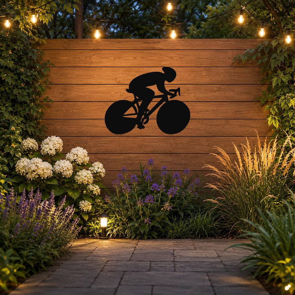 Black metal wall art featuring a bold silhouette of a cyclist on a road bike, mounted on a wooden outdoor wall above a garden with string lights and plants.
