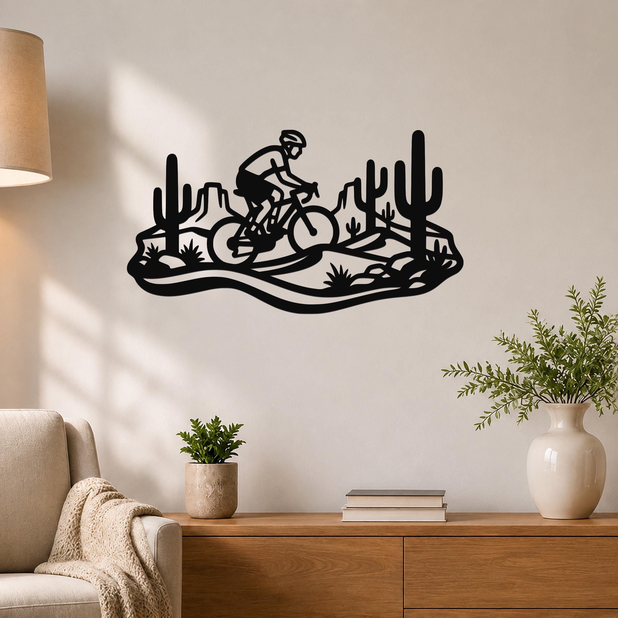 Black metal wall art silhouette of a cyclist on a mountain bike through a Sonoran desert landscape with saguaro cacti, mounted on a living room wall above a wooden console table.