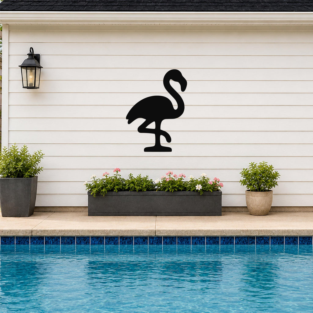 Black metal flamingo wall art silhouette mounted on a white siding wall above a poolside planter with flowers and potted plants.