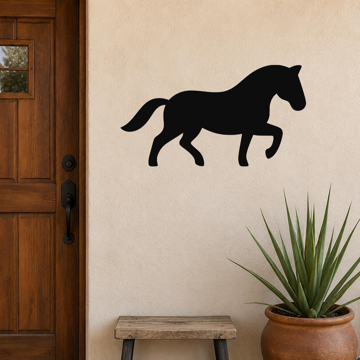 Black metal horse silhouette wall art mounted on a beige stucco wall beside a wooden door, with a potted plant and small bench below.