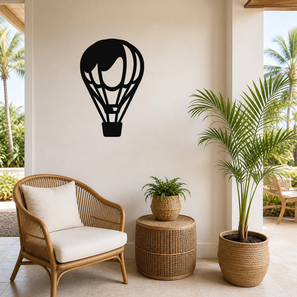 Black metal hot air balloon wall art with internal basket and panel cutouts, mounted on a light-colored wall above a wicker chair in a tropical patio setting.