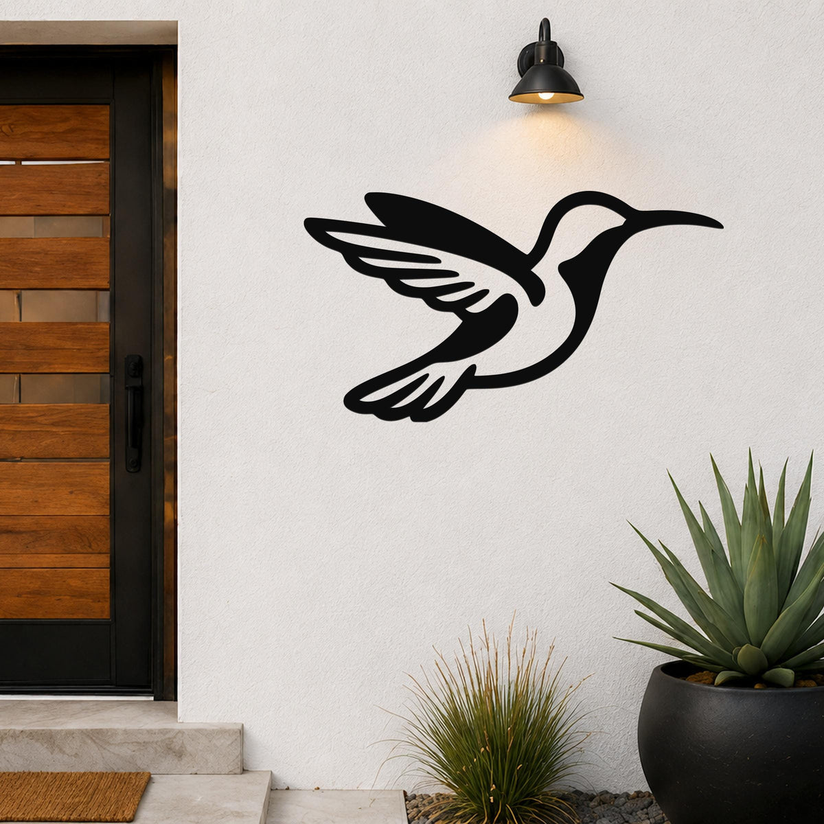 Black metal hummingbird wall art with wing and tail cutouts, mounted on a white exterior wall beside a wooden door and potted plants.