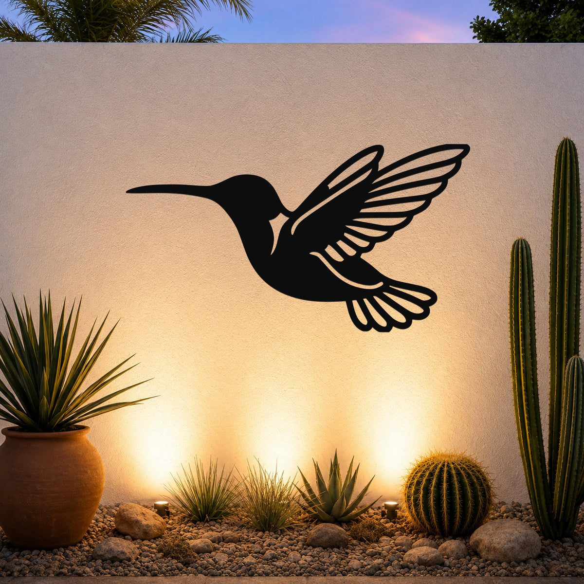 Black metal hummingbird wall art with wing and tail cutouts, mounted on an outdoor wall above a desert garden with cacti and warm uplighting.