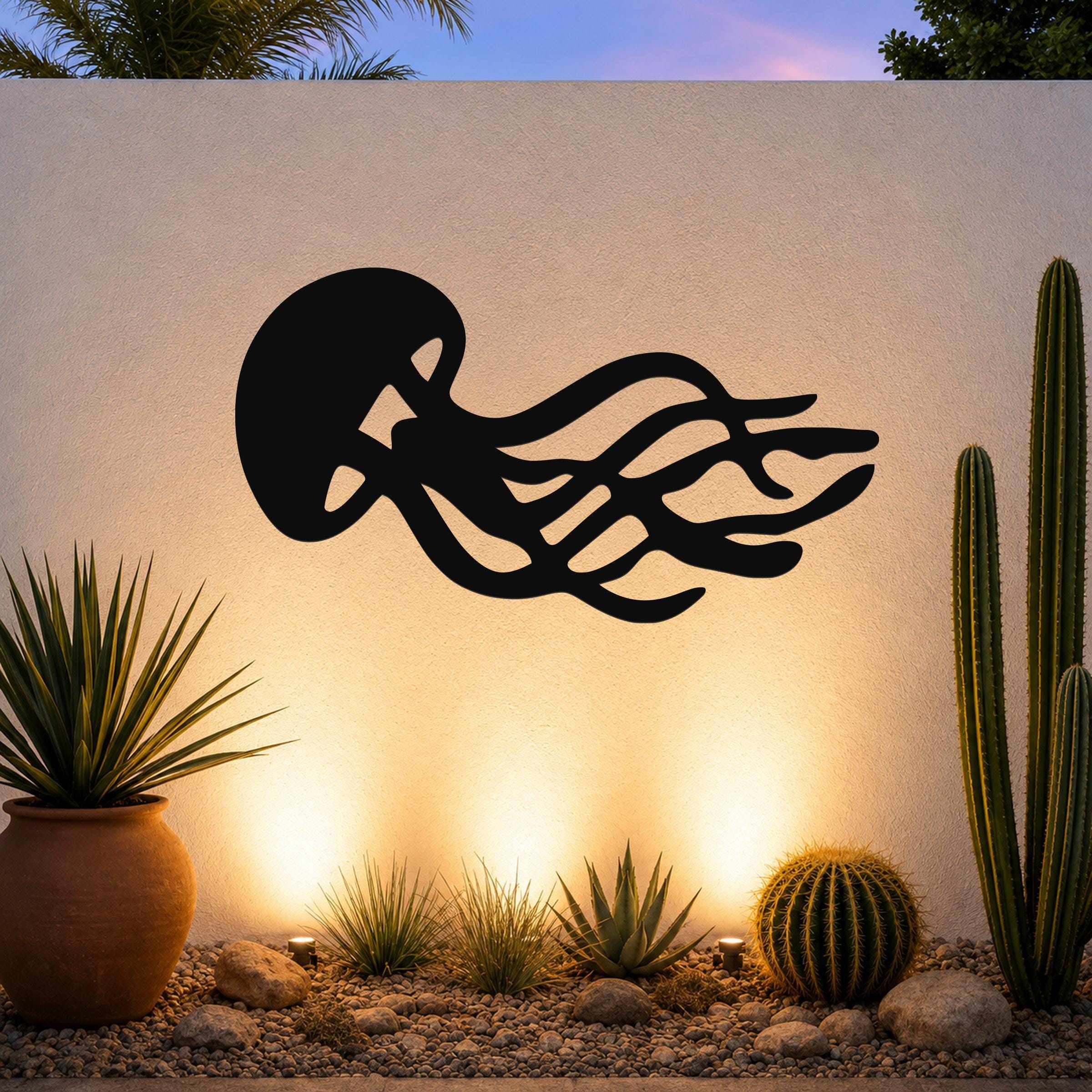 Black metal jellyfish wall art with flowing tentacle cutouts, mounted on an outdoor wall above a desert garden with cacti and warm uplighting.
