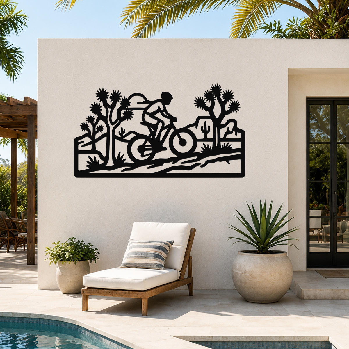 Black metal wall art cutout of a cyclist on a desert trail with Joshua trees and cacti, mounted on an outdoor patio wall above a lounge chair.