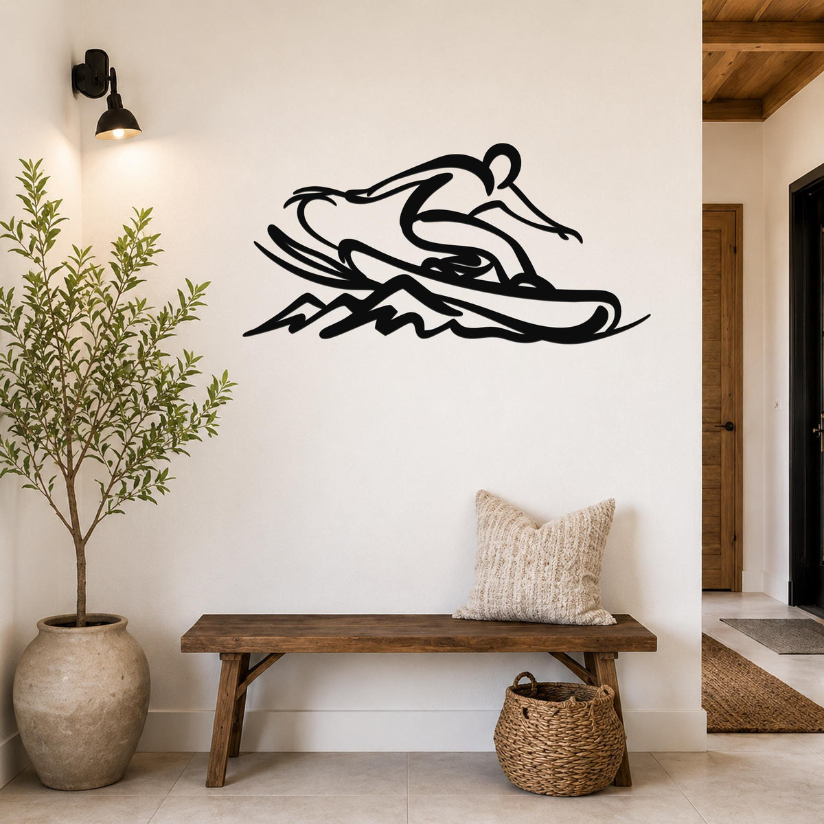 Black metal wall art silhouette of a snowboarder in motion, mounted on a white wall above a wooden bench in a hallway.