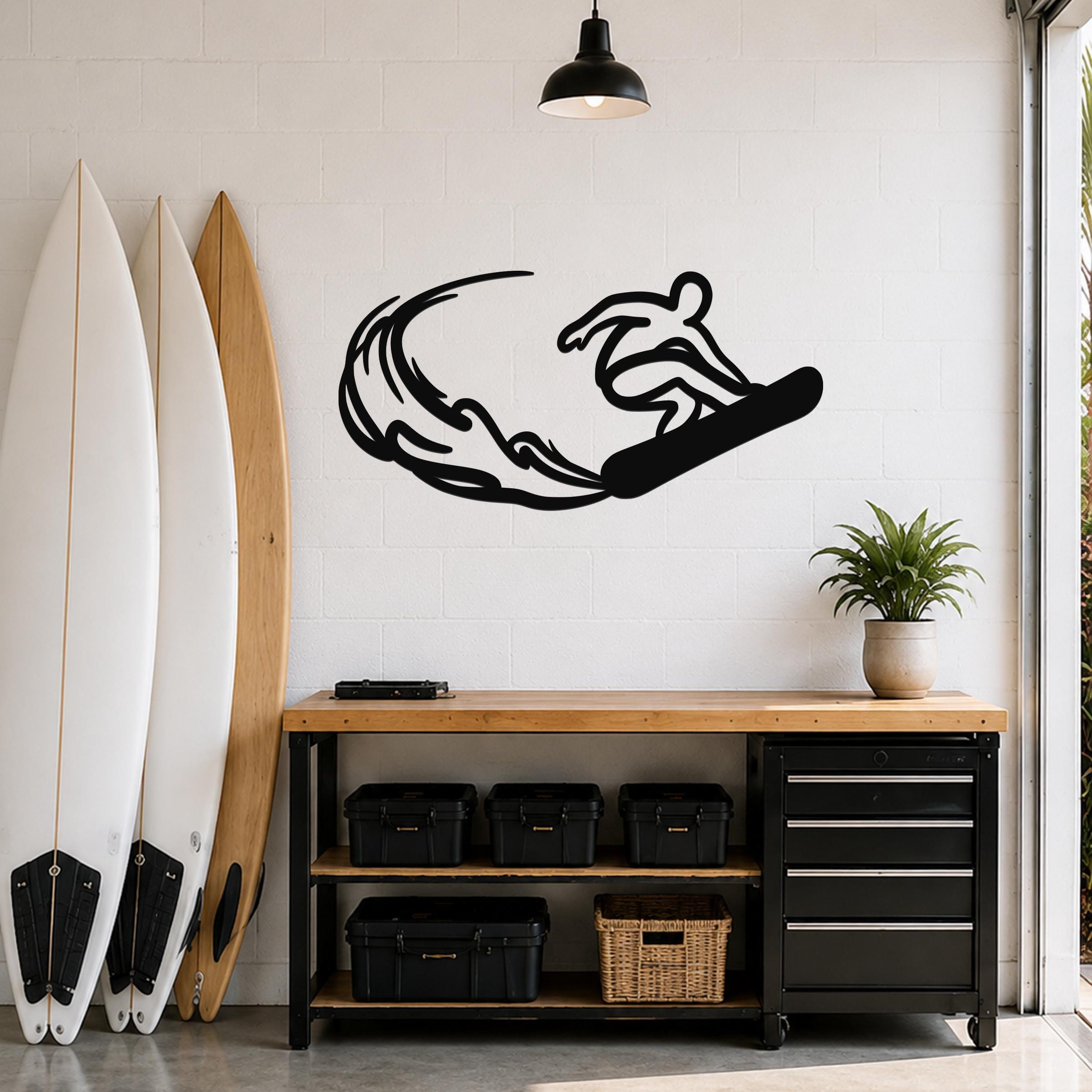 Black metal wall art silhouette of a snowboarder in motion with wave-like cutouts, mounted on a white brick wall above a wooden workbench in a garage.