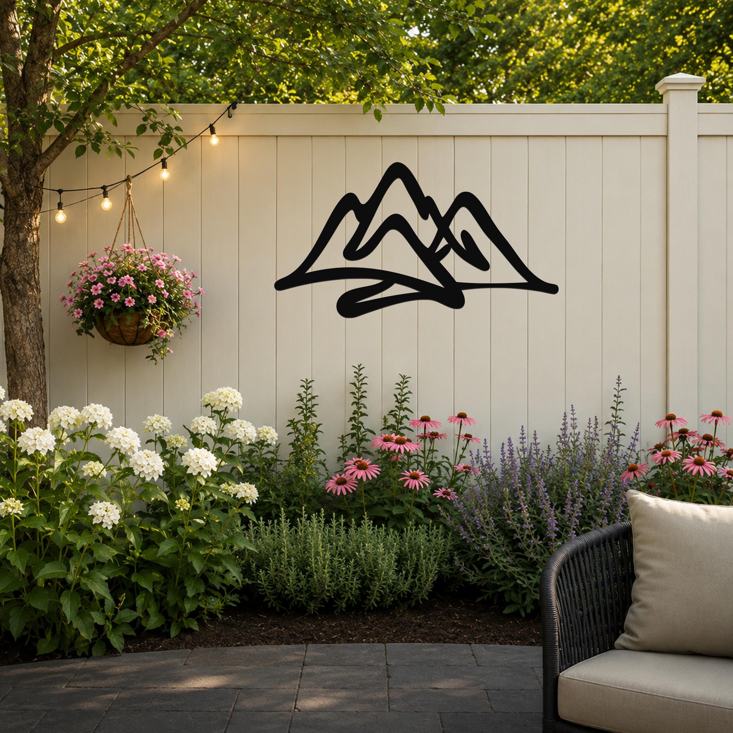 Black metal mountain silhouette wall art mounted on a white outdoor fence, above a garden bed with flowers and string lights.