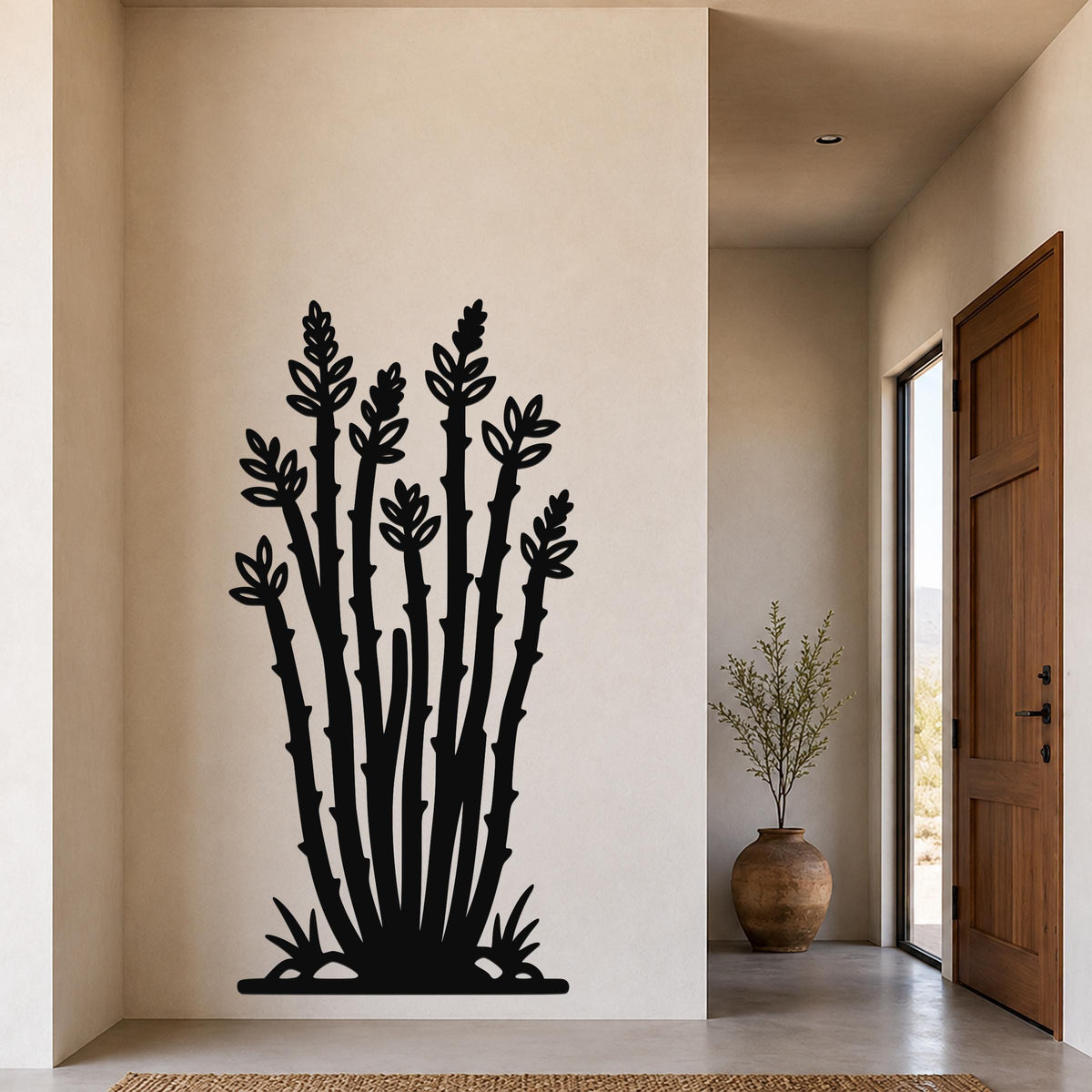 Black metal ocotillo wall art with leafy branch cutouts, mounted on a beige wall in a hallway near a wooden door and large vase.