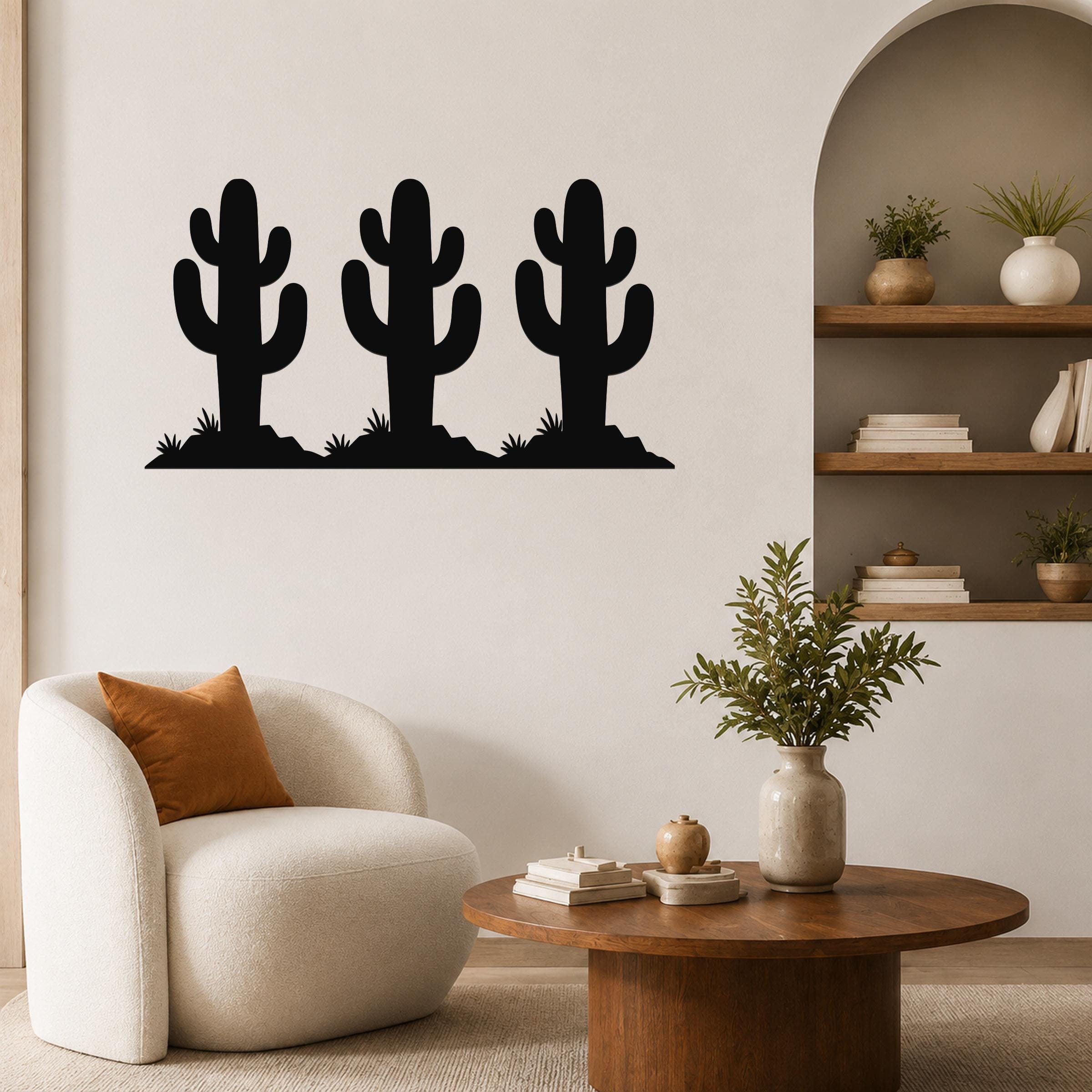 Black metal wall art featuring a trio of saguaro cactus silhouettes with spines and base grass, mounted on a living room wall above a white armchair.