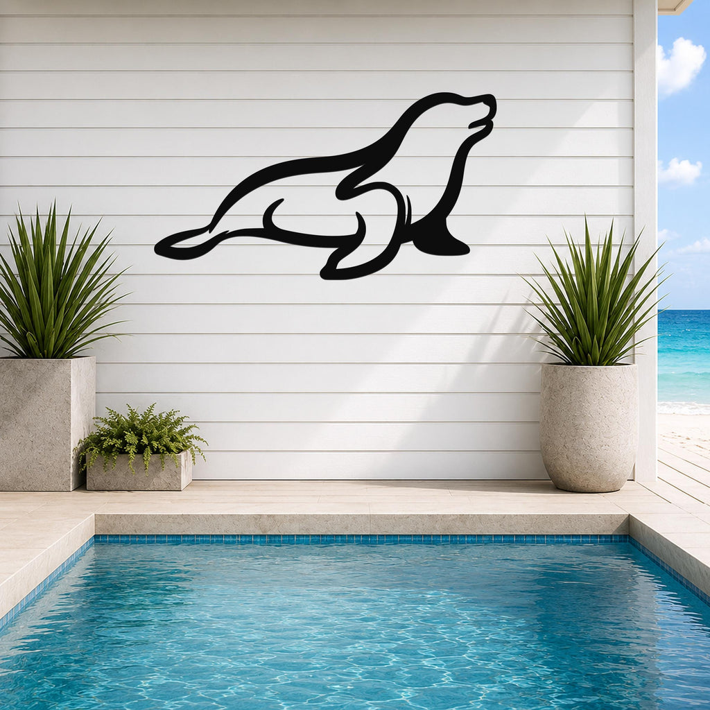 Black metal sea lion wall art with a sleek silhouette design, mounted on a white shiplap wall above a pool with potted plants and ocean views.