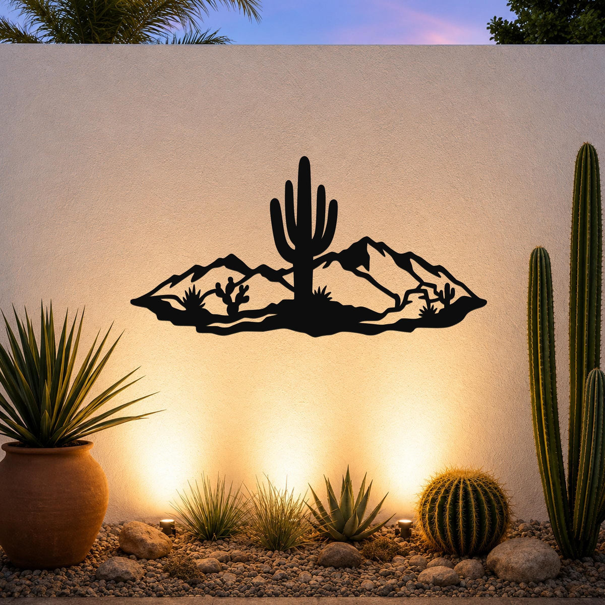 Black metal wall art silhouette of a desert scene with a saguaro cactus and mountains, mounted on an outdoor wall above a gravel garden with cacti and warm uplighting.