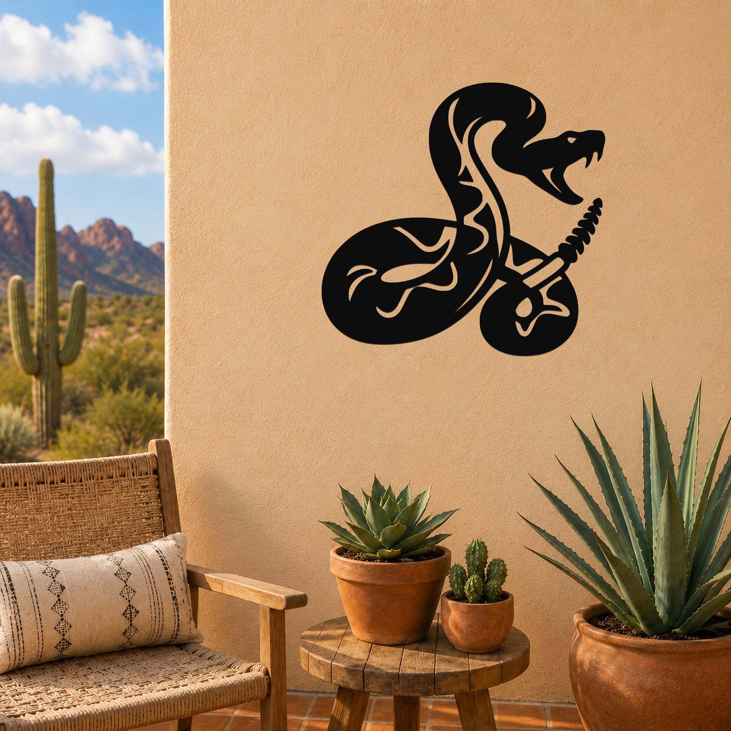Black metal wall art featuring a coiled rattlesnake silhouette with internal cutout details, mounted on a tan outdoor wall above a small wooden table with potted succulents.