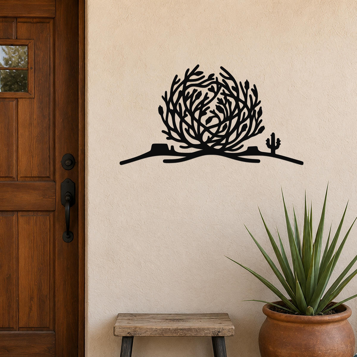 Black metal wall art cutout of a tumbleweed and cactus on a desert hill, mounted on a beige exterior wall beside a wooden door and potted plant.