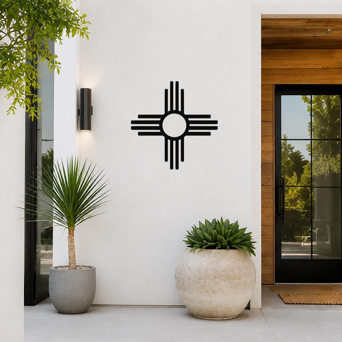 Black metal Zia Sun symbol wall art with a bold circular design and radiating lines, mounted on a white exterior wall beside a glass door and potted plants.