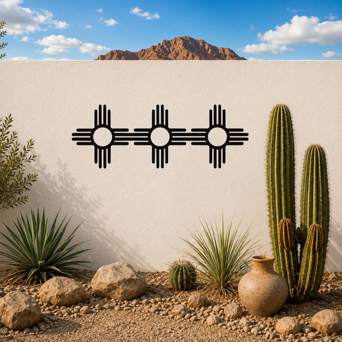 Black metal Zia Sun symbol trio wall art with four rays and central circles, mounted on a white outdoor wall above a desert garden with cacti and rocks.