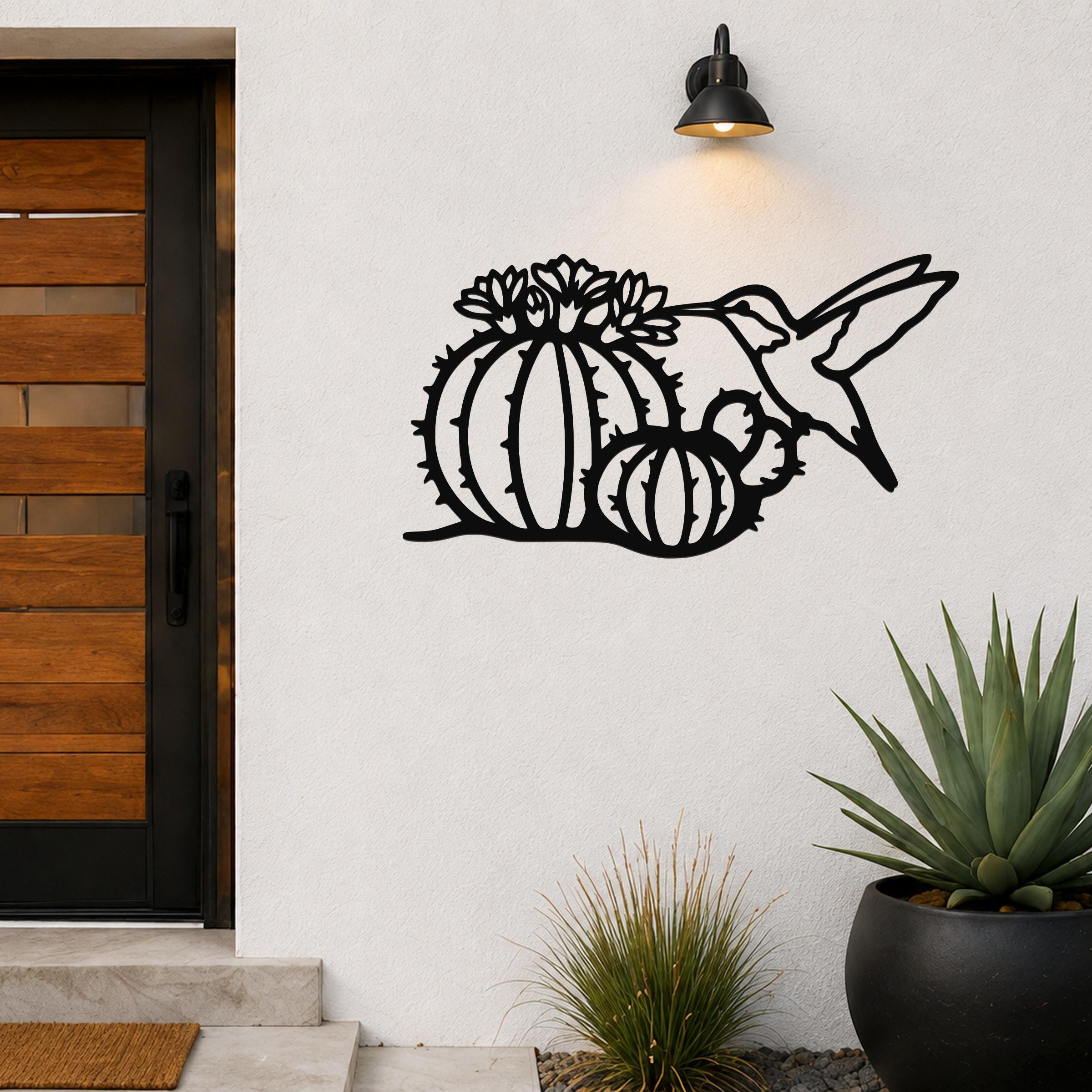 Black metal wall art featuring a hummingbird and barrel cactus with flower cutouts, mounted on a white exterior wall beside a wooden door.