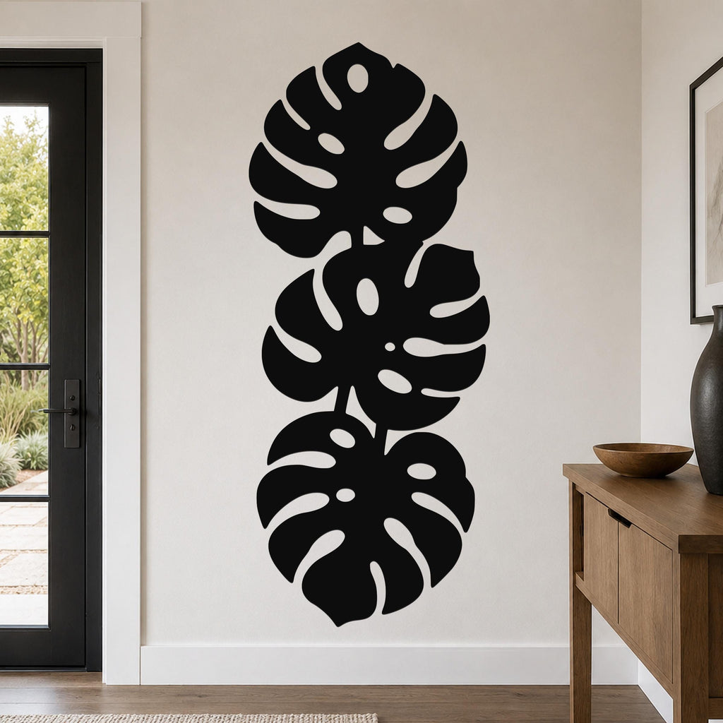Black metal wall art featuring three stacked monstera leaf cutouts, mounted vertically on a living room wall above a wooden console table.