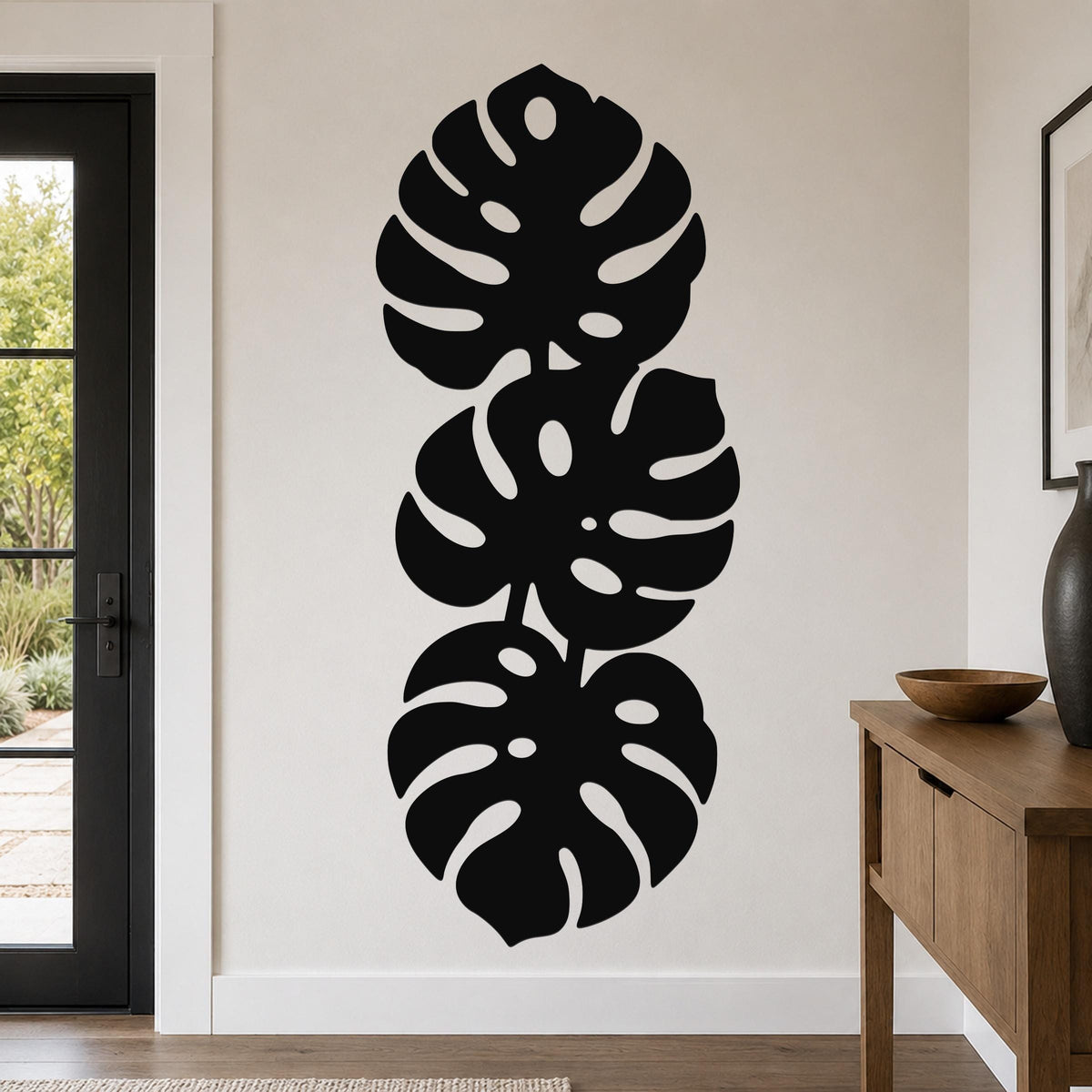 Black metal wall art featuring three stacked monstera leaf cutouts, mounted vertically on a living room wall above a wooden console table.