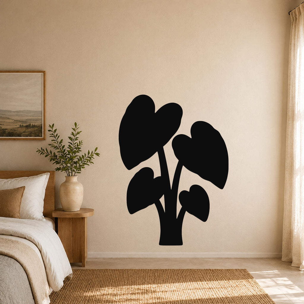 Black metal wall art in the shape of an elephant ear plant with large leaf cutouts, mounted on a beige bedroom wall above a woven rug.