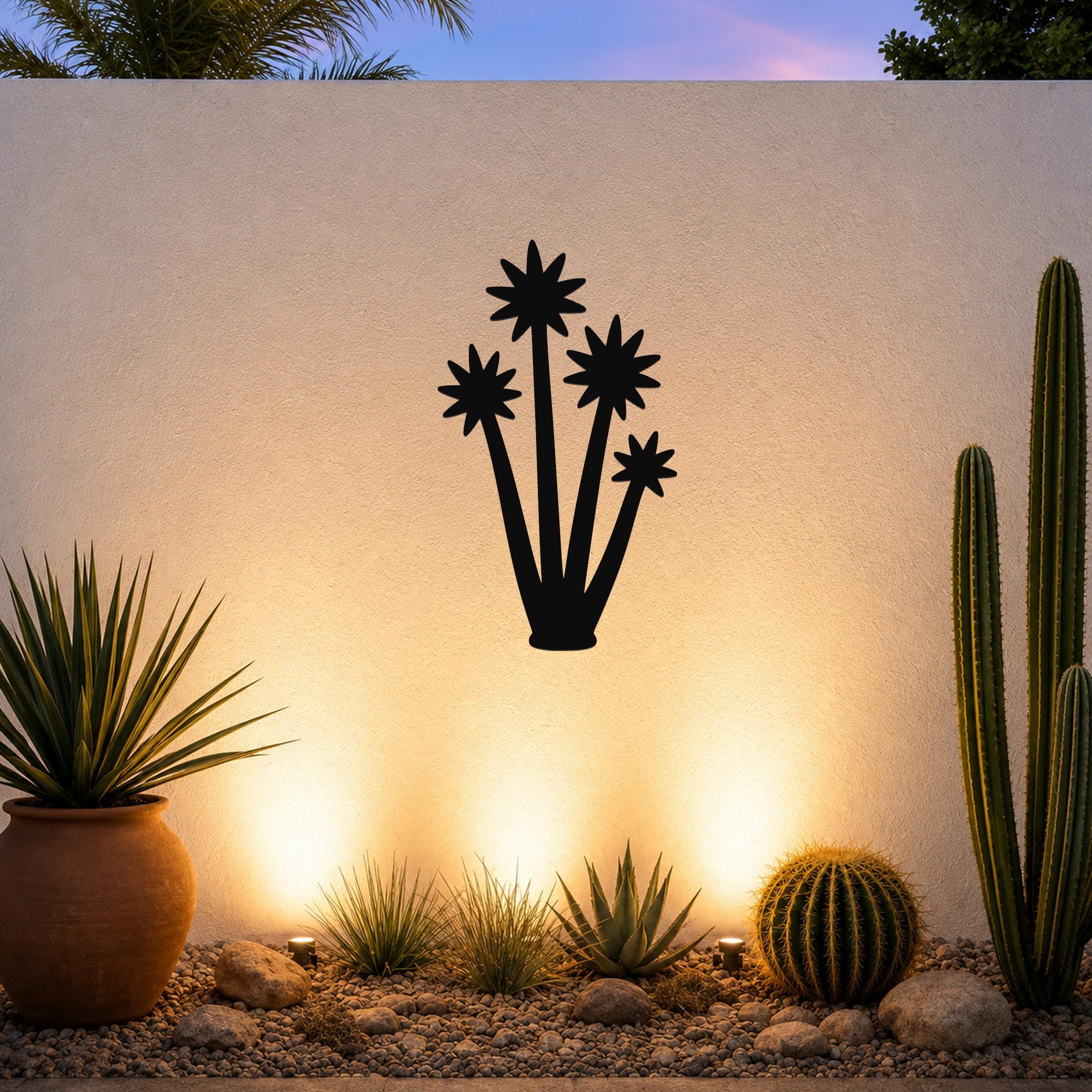 Black metal wall art silhouette of a tall yucca plant with star-shaped blooms, mounted on a textured outdoor wall above a desert garden with potted succulents and ground lights.