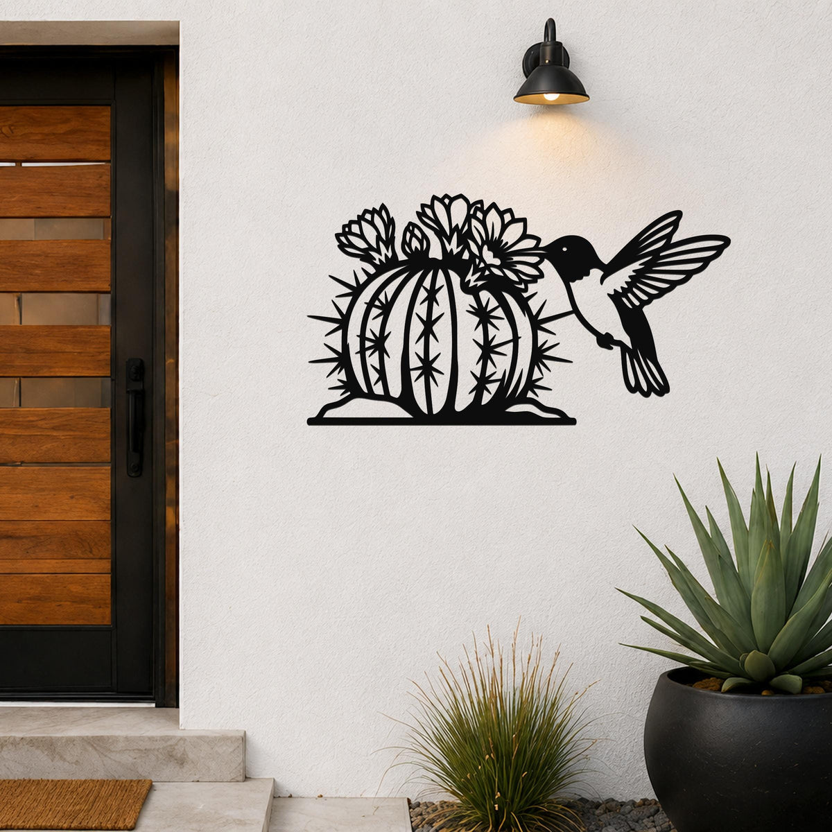 Black metal wall art featuring a hummingbird and flowering barrel cactus cutout, mounted on a white exterior wall beside a front door.