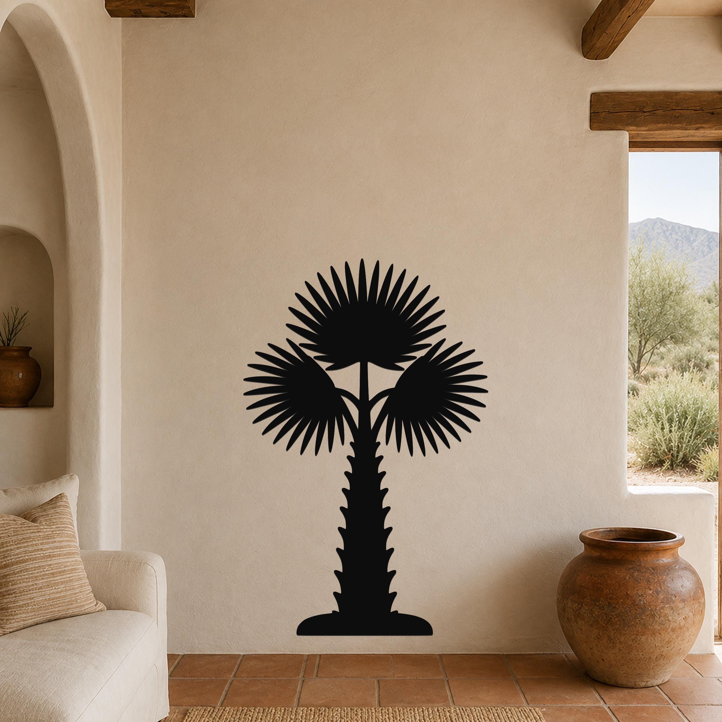 Black metal palm tree wall art with fan-shaped fronds and a textured trunk, mounted on a beige stucco wall in a living room with terracotta flooring and wooden beams.