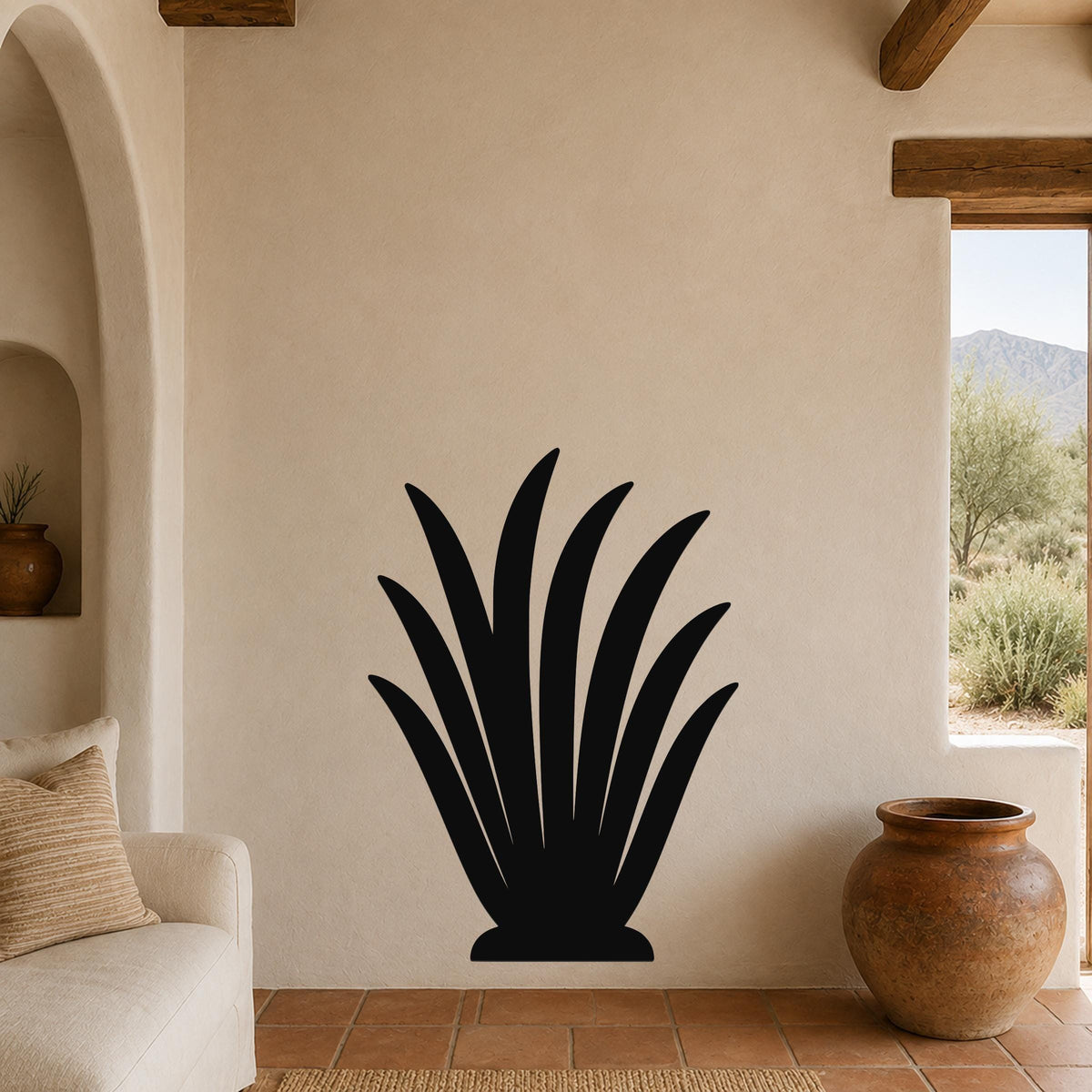 Black metal agave plant wall art with leaf cutouts, mounted on a beige stucco wall in a living room with terracotta flooring and wooden beams.