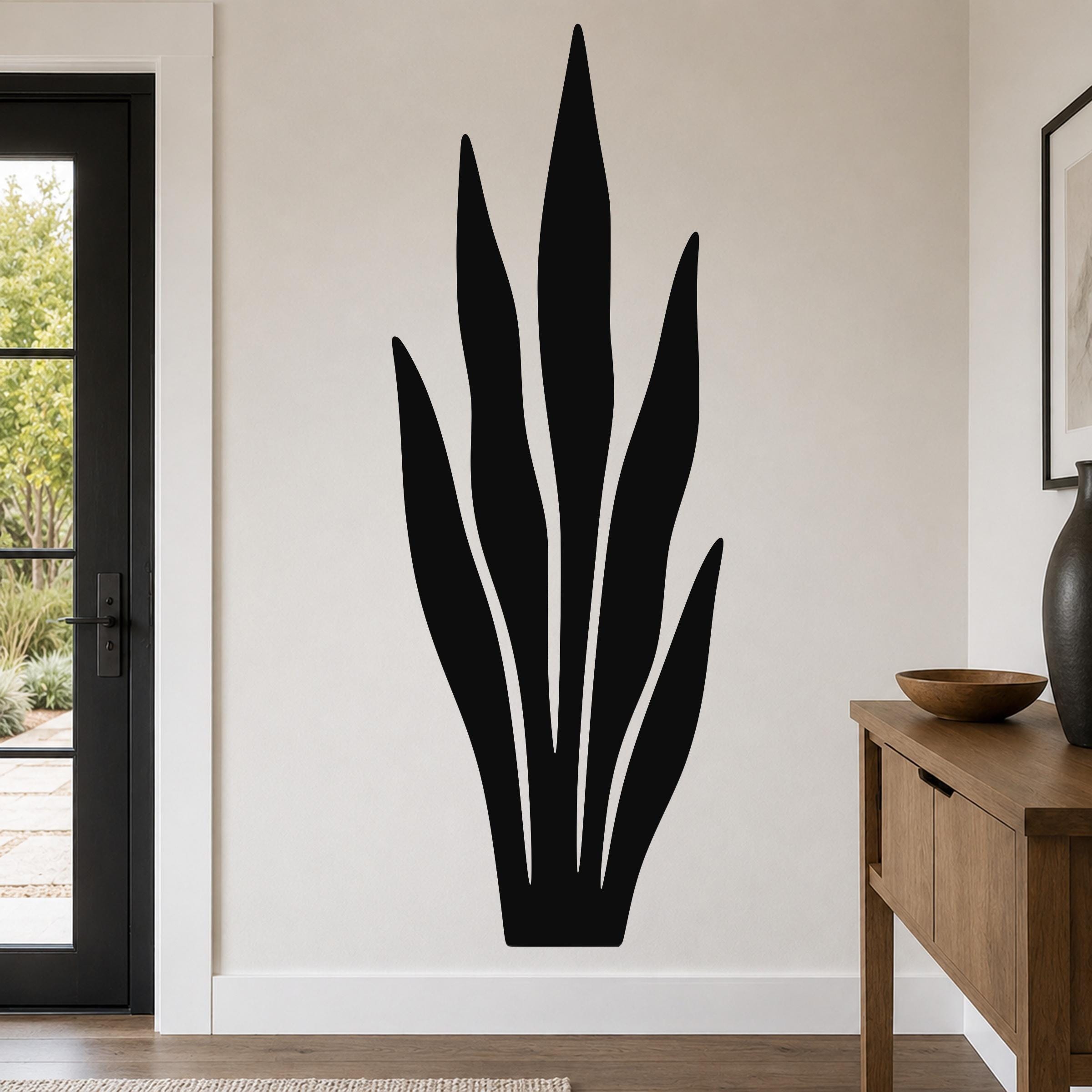 Black metal wall art in a tall, leafy plant silhouette with internal cutouts, mounted on a light-colored wall above a wooden console table in a modern entryway.