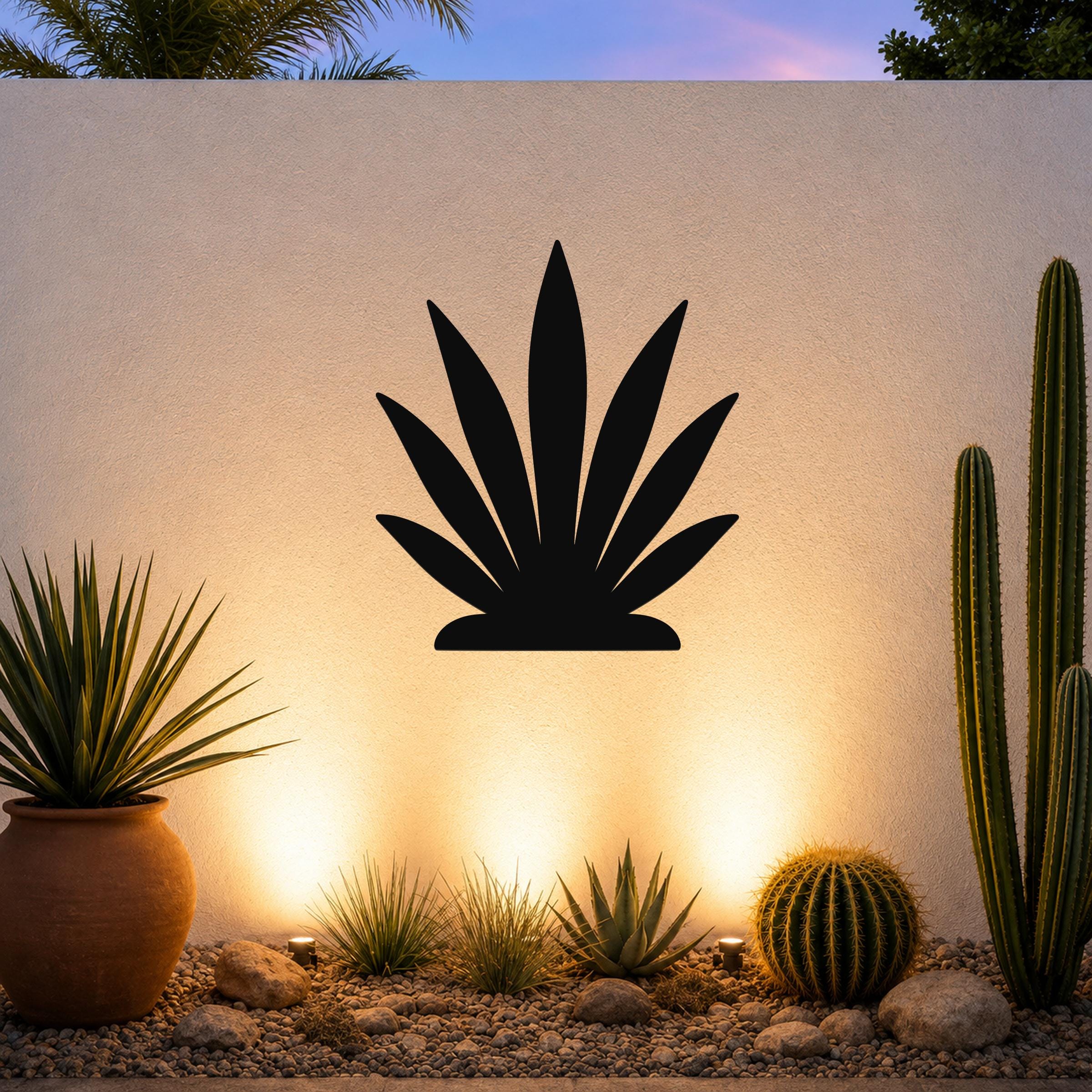 Black metal agave wall art silhouette mounted on a textured outdoor wall above a desert garden with cacti and warm uplighting.