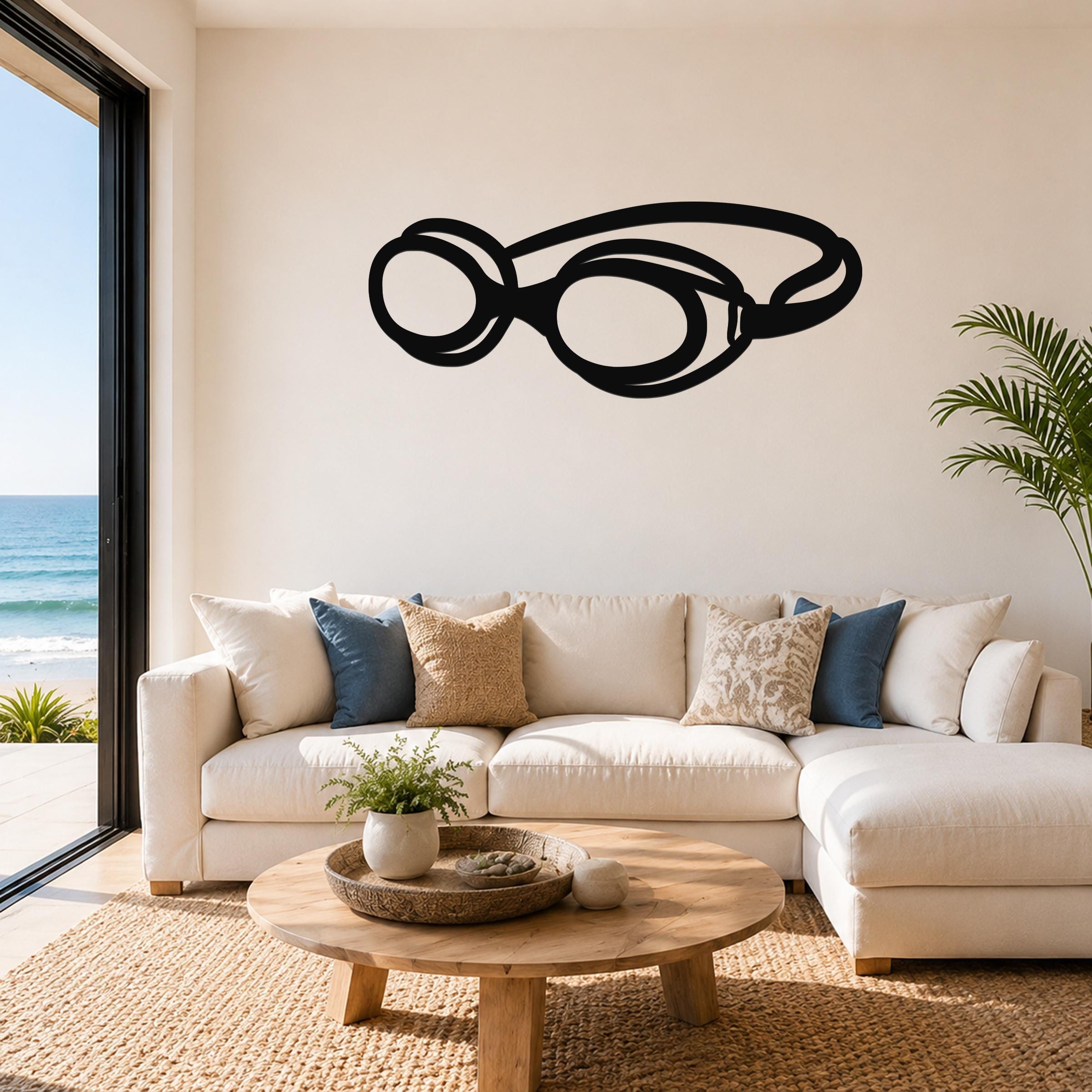 Black metal swim goggles wall art with cutout design, mounted above a white sofa in a living room with ocean views.