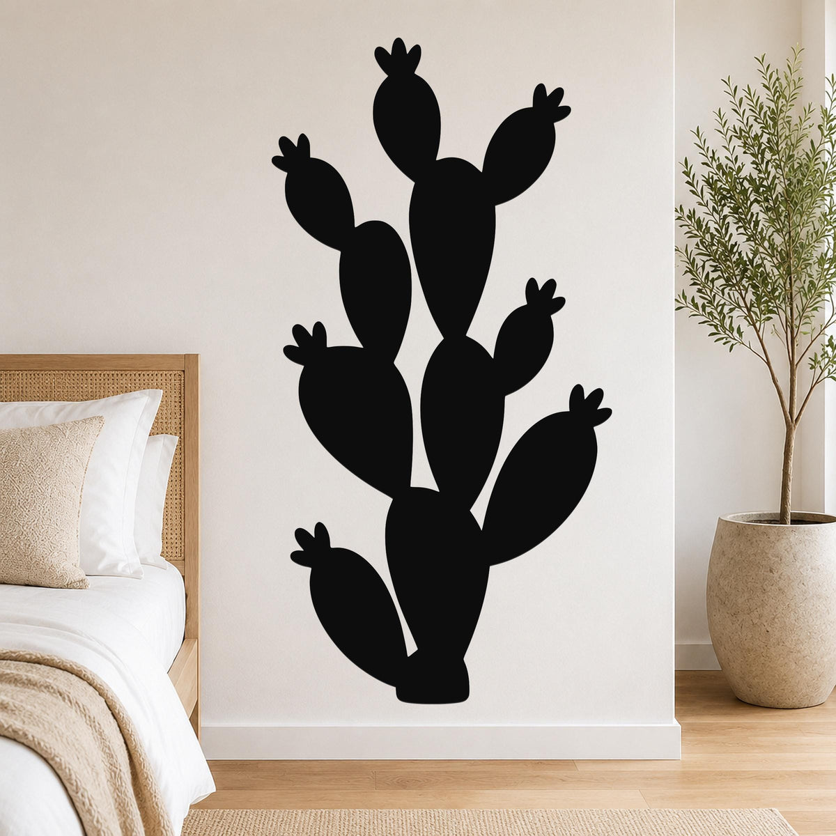 Black metal cactus wall art with multiple paddle-shaped segments and small flower cutouts, mounted on a white bedroom wall above a bed.