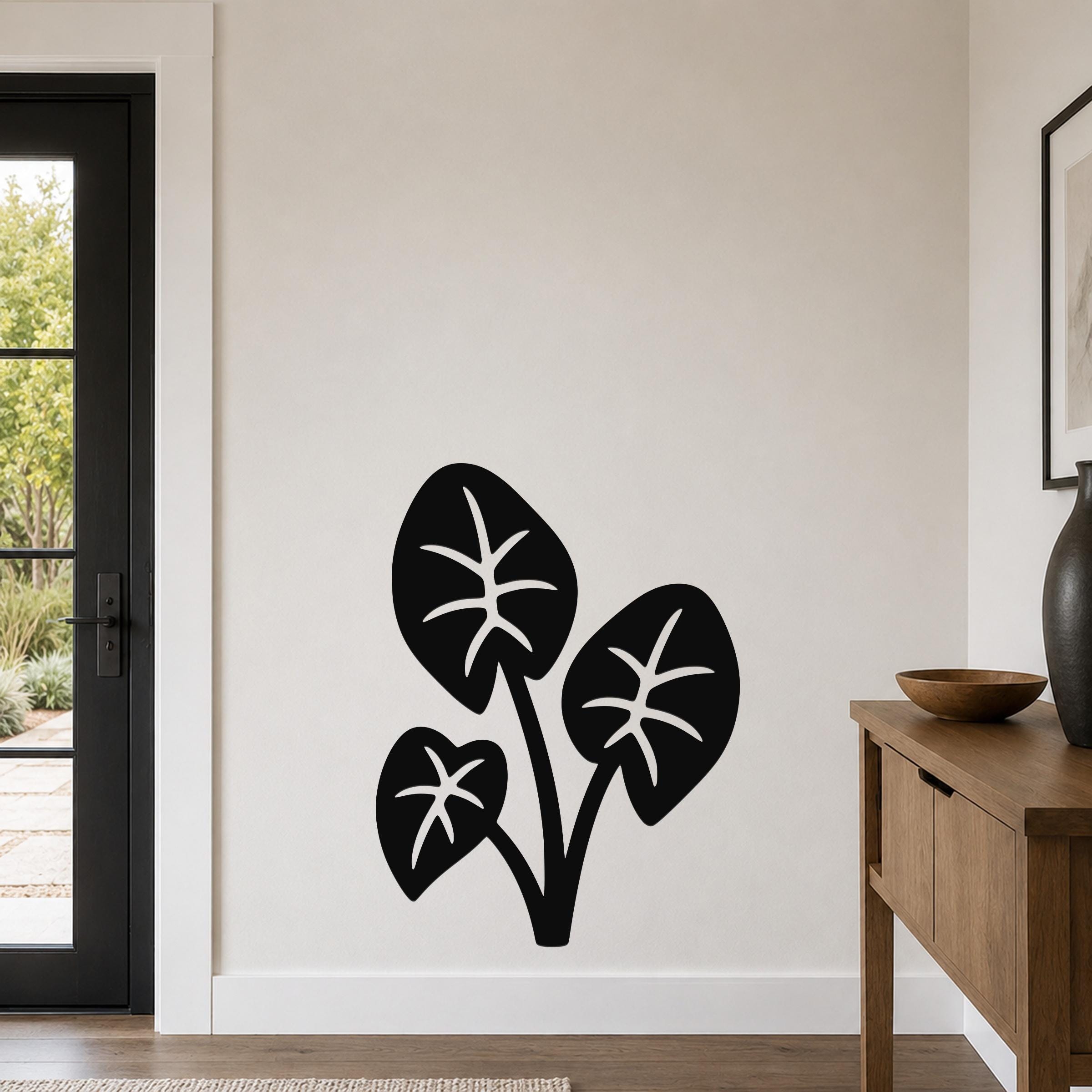Black metal wall art featuring a cutout silhouette of three elephant ear leaves, mounted on a light-colored wall in a modern entryway.