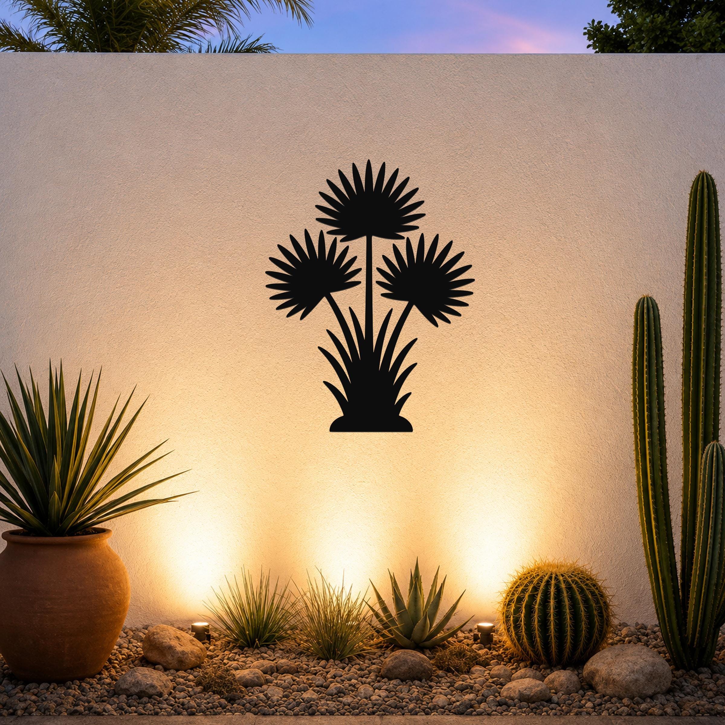 Black metal fan palm tree wall art with three frond silhouettes, mounted on a white outdoor wall above a desert garden with cacti and warm uplighting.