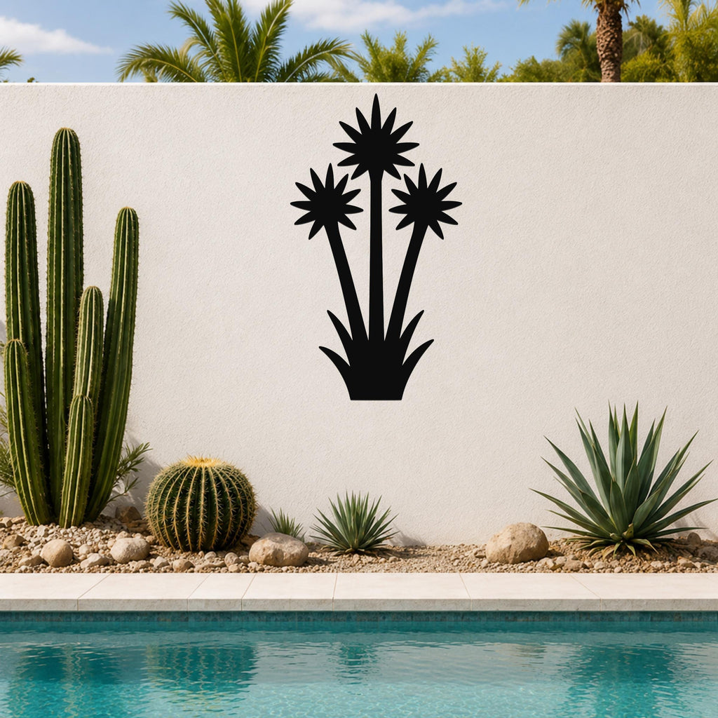 Black metal wall art cutout of a blooming yucca plant with three starburst flowers and spiky base, mounted on a white outdoor wall above a poolside garden.