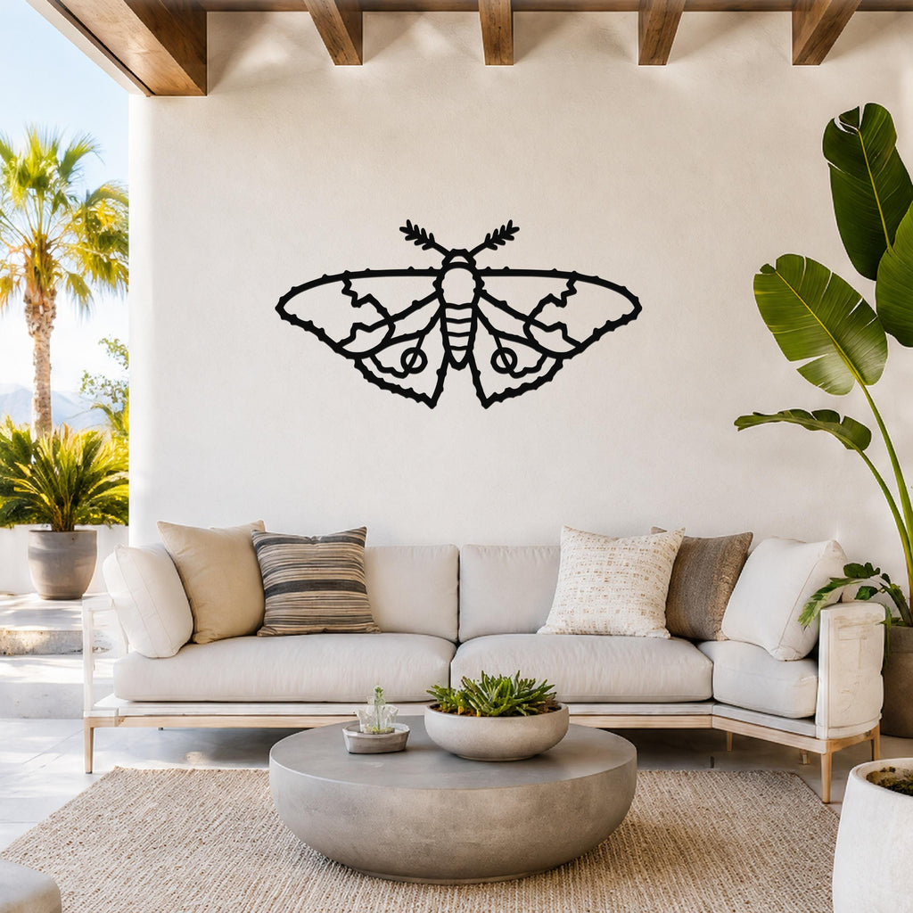 Black metal moth wall art with detailed wing cutouts, mounted on a white wall above a light-colored sofa in a bright living room.