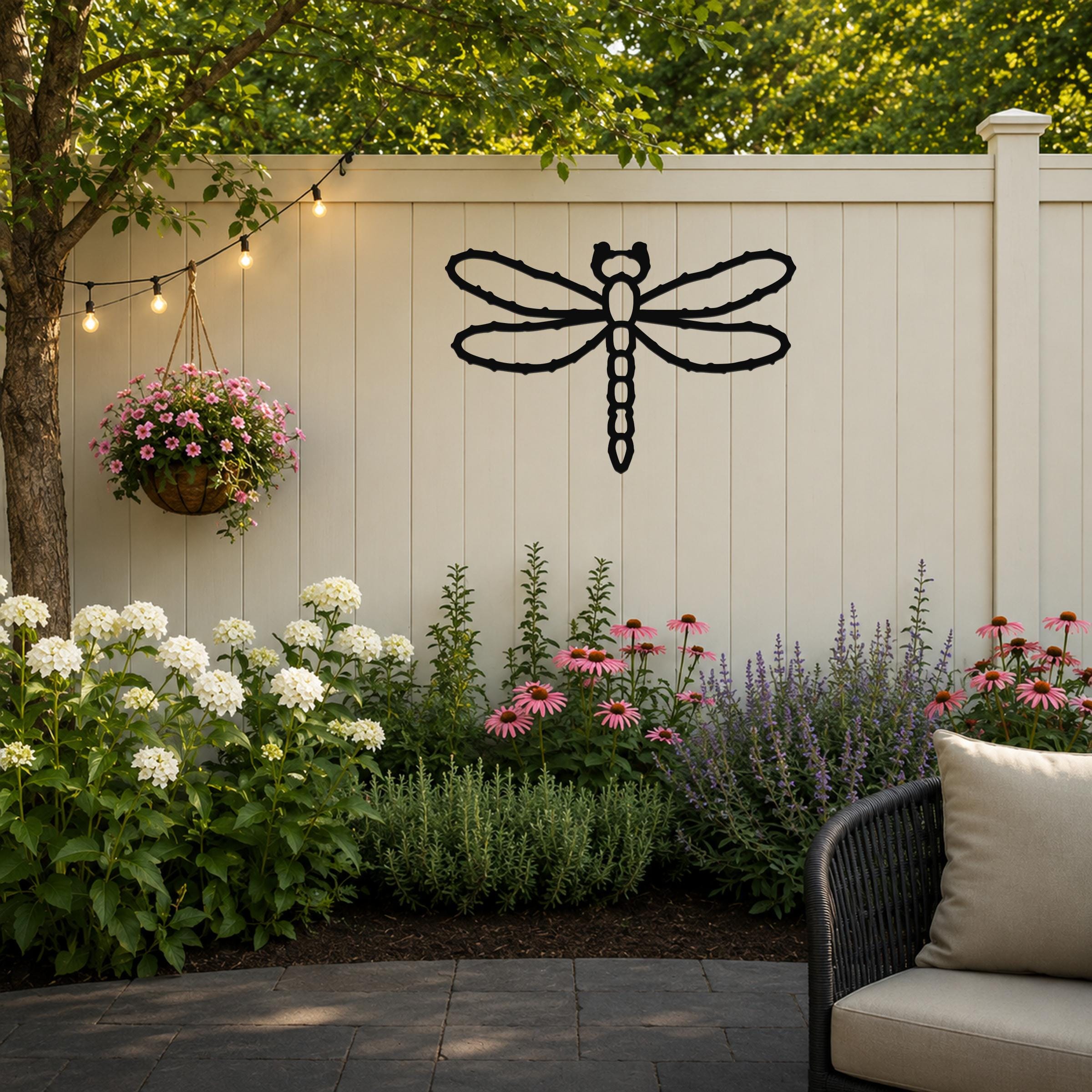 Black metal dragonfly wall art with detailed wing cutouts, mounted on a light beige outdoor fence above a flower bed.