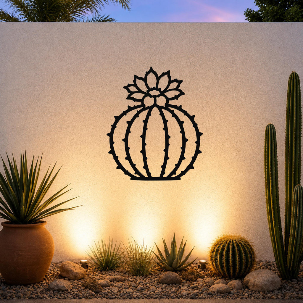 Black metal barrel cactus wall art with flower and spine cutouts, mounted on a white outdoor wall above a desert garden with potted plants and ground lights.